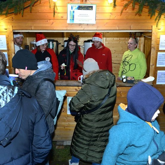 Eine Gruppe von Menschen steht vor einem kleinen Holzhäuschen. Zwei Frauen stehen hinter der Theke. Der Mann rechts trägt einen Weihnachtsmannhut.