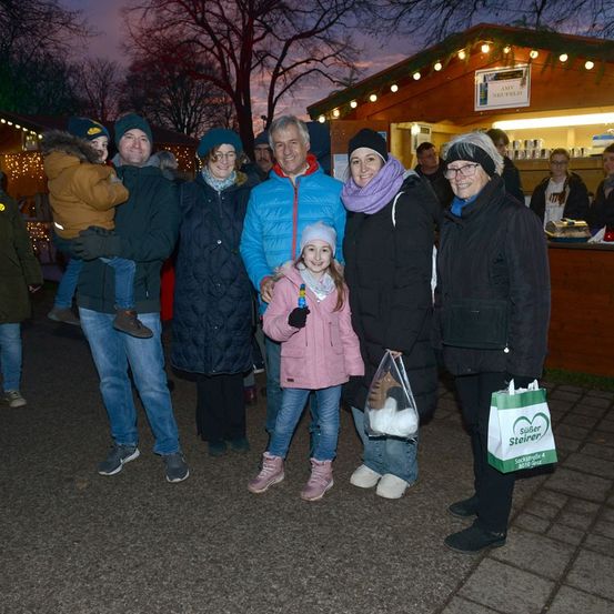 Eine Familie mit fünf Personen steht in einer festlichen Umgebung im Freien, mit Winterkleidung und einem Kind, das ein Plüschtier hält. Dahinter lächeln zwei Frauen, und eine hält eine Tasche mit einem Logo. Im Hintergrund ist ein Stand mit Dekorationen und einem Schild zu sehen.
