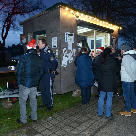 Ein kleines Gebäude mit Lichtern und einem Polizisten im Inneren, Leute stehen darum herum, einer hält einen Becher, ein anderer trägt einen Weihnachtsmannhut und eine Frau trägt eine Brille.
