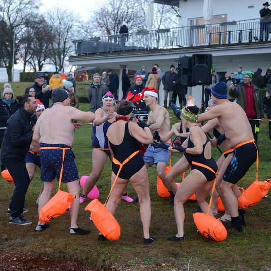 Eine Gruppe von Menschen, die in Badekleidung und Weihnachtsmützen gekleidet sind und orangefarbene Taschen halten, tanzt auf einem Rasen, während andere hinter einer Barriere zusehen.