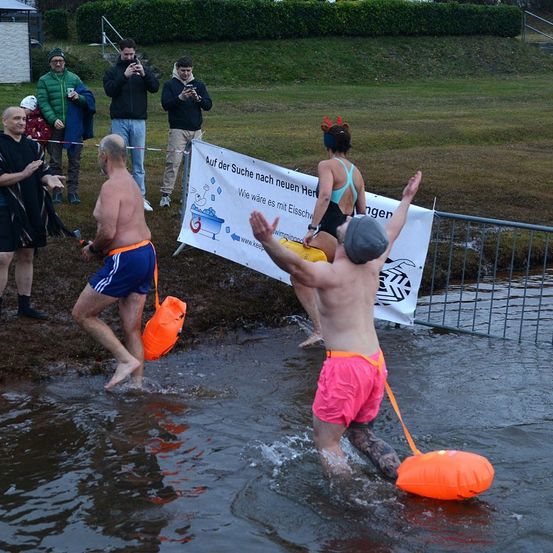 Mehrere Personen waten durch ein Gewässer, wahrscheinlich einen Fluss, in farbenfroher Badekleidung. Ein Mann trägt pinkfarbene Shorts, ein anderer eine graue Mütze und orangefarbene Schwimmhilfen. Dahinter befindet sich ein Banner mit Text und Logo.