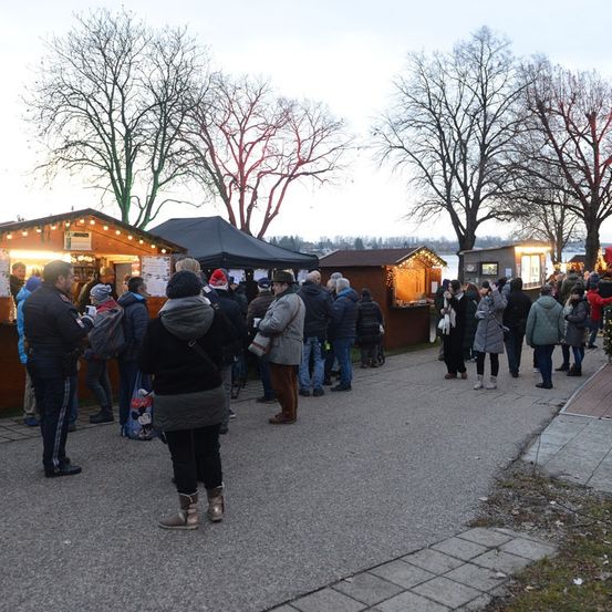 Eine Gruppe von Menschen steht draußen auf einem Weg, gesäumt von kleinen Geschäften und Bäumen, unter einem grauen Himmel. Einige tragen Mützen und Winterkleidung.