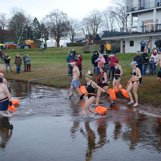 Eine Gruppe von Menschen, einige in Badekleidung, durchquert einen Fluss und trägt orangefarbene Eimer. Zuschauer beobachten vom Grasufer aus, mit einem Gebäude im Hintergrund.