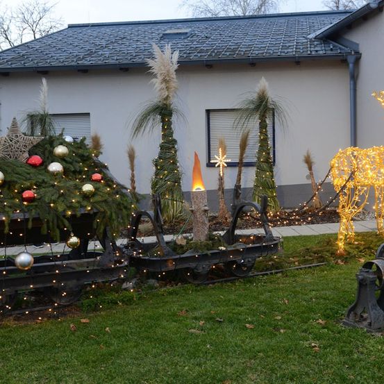 Ein festlicher Weihnachtsschlitten mit Dekorationen steht vor einem Haus mit grauem Dach. Der Schlitten ist mit Weihnachtskugeln und Lichtern geschmückt.