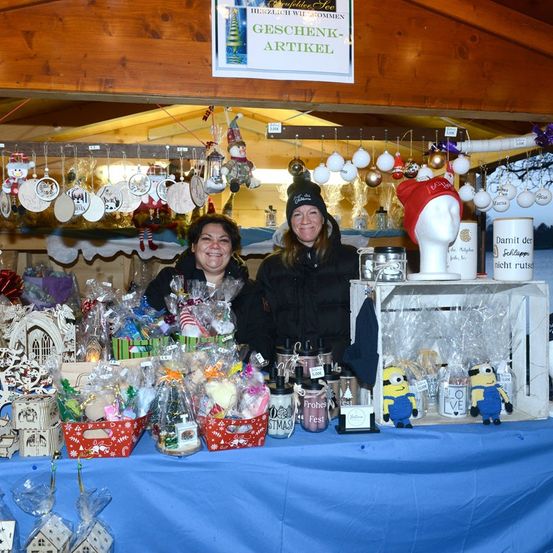 Zwei Frauen stehen hinter einem Tisch, auf dem verschiedene Weihnachtsgeschenke wie Körbe, Tassen und Figuren zu sehen sind, unter einem Schild, das Geschenk Artikel steht.