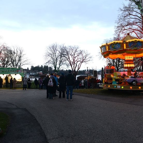 Eine Menschenmenge versammelt sich auf einem Freiluft-Jahrmarkt mit einem Karussell im Vordergrund und einem Imbisswagen auf der linken Seite. Die Szene spielt in der Dämmerung, mit Bäumen und einem Gewässer im Hintergrund.