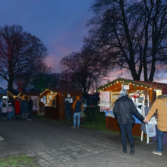 Eine Gruppe von Menschen versammelt sich um Stände auf einem Nachtmarkt, beleuchtet von Weihnachtslichtern. Ein Paar geht Hand in Hand an einem der Stände vorbei.