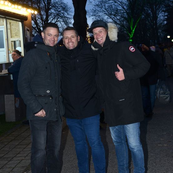 Drei Männer stehen lächelnd bei Nacht in einem festlichen Außenbereich mit beleuchteten Bäumen und einer verschwommenen Menschenmenge im Hintergrund.