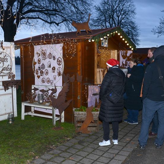 Eine Gruppe von Menschen in Winterkleidung besucht einen festlichen Weihnachtsstand, der mit Lichtern, Ornamenten und einer hölzernen Katze auf dem Dach dekoriert ist.