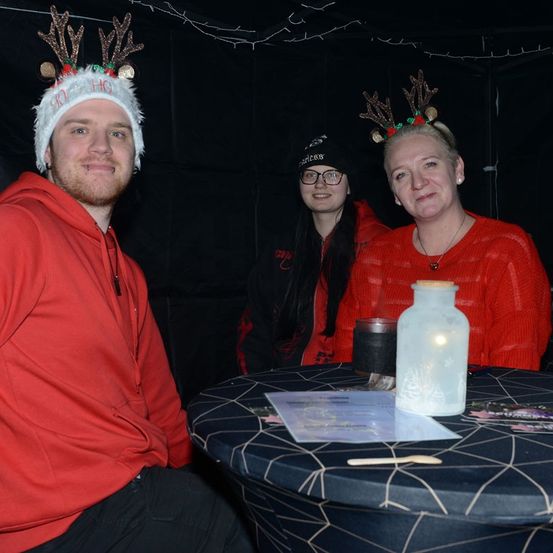 Drei Menschen sitzen in einem schwach beleuchteten Raum, vielleicht für eine Weihnachtsveranstaltung. Ein Mann und eine Frau tragen Weihnachtsmützen. Vor ihnen steht ein runder Tisch mit Papieren, einer Flasche und einer Kerze.