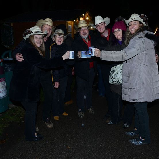 Eine Gruppe von Menschen, die Cowboyhüte tragen, lächeln und halten Becher in einer nächtlichen Umgebung. Sie scheinen für ein Foto zu posieren.
