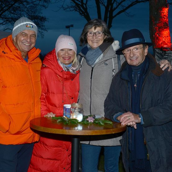 Vier Erwachsene stehen nachts lächelnd zusammen. Sie sind warm gekleidet, zwei von ihnen tragen Mützen. Vor ihnen steht ein kleiner Tisch mit einer Kerze und einem Kranz. Sie posieren wahrscheinlich für ein Foto.