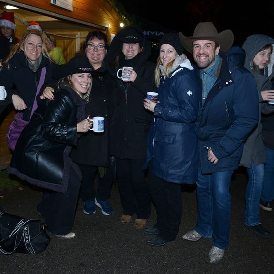 Eine Gruppe von Menschen in Winterkleidung posiert bei Nacht für ein Foto und hält Kaffeebecher in der Hand. Sie scheinen sich auf einem Weihnachtsmarkt zu befinden.