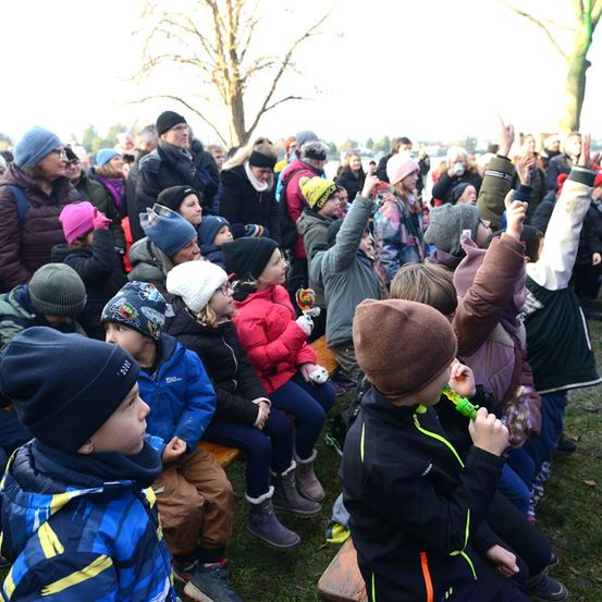 Eine große Gruppe von Menschen, einschließlich Kinder, versammelt sich im Freien auf einem Grasbereich, wahrscheinlich in einem Park. Sie tragen Winterkleidung, und einige halten ihre Hände hoch. Sie beobachten eine Veranstaltung, und einige halten Spielzeug.