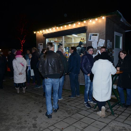 Eine Gruppe von Menschen steht bei Nacht vor einem Essensstand, der von festlichen Lichtern beleuchtet wird. Einige kaufen Essen, während andere in der Schlange warten.