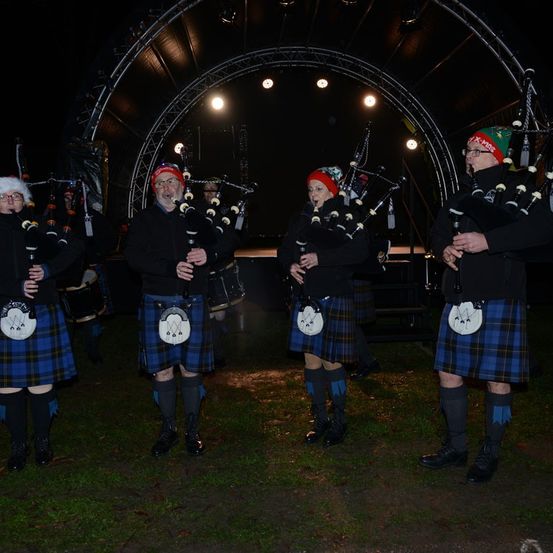 Eine Gruppe von Dudelsackspielern in Kilts tritt bei Nacht auf einer Bühne auf. Sie spielen ihre Instrumente unter einer Reihe von Scheinwerfern.