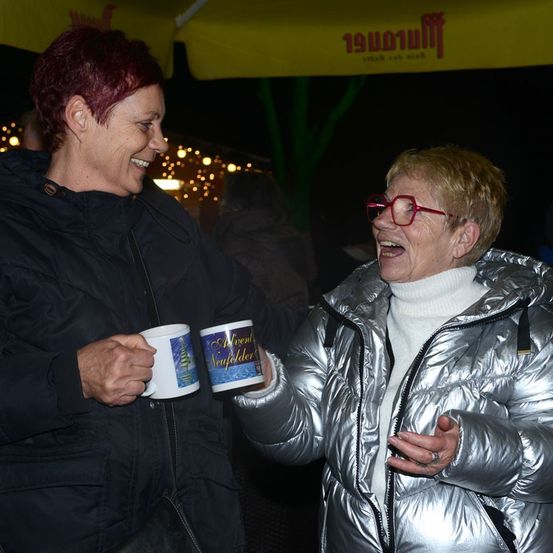 Zwei Frauen lachen und stoßen mit Tassen bei einem Weihnachtsmarkt im Freien an, beide in festlicher Kleidung. Eine hält eine Tasse mit 'Advent Neufelder' Text, und sie stehen unter einem gelben Zelt.