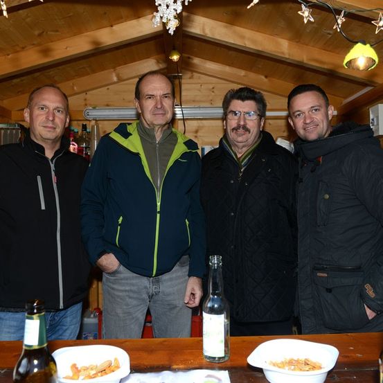 Vier Männer stehen in einer Hütte. Sie lächeln und scheinen für ein Foto zu posieren. Flaschen und Schüsseln mit Essen stehen auf einem Tisch vor ihnen.