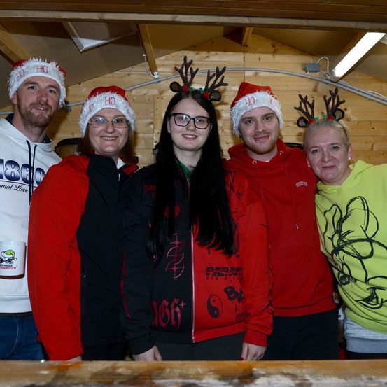 Fünf Menschen stehen eng beieinander und tragen Weihnachtsmützen mit Rentiergeweihen. Sie lächeln für ein Foto in einem hölzernen Raum.