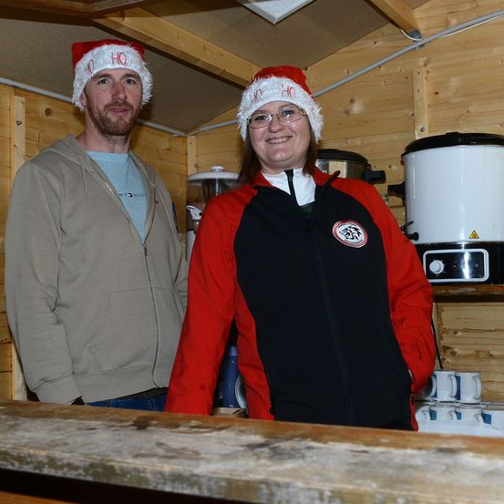 Ein Mann und eine Frau mit Weihnachtsmützen stehen nebeneinander in einer kleinen Holzhütte und lächeln für ein Foto. Sie sind von verschiedenen Gegenständen umgeben, darunter Kaffeetassen und ein Reiskocher.