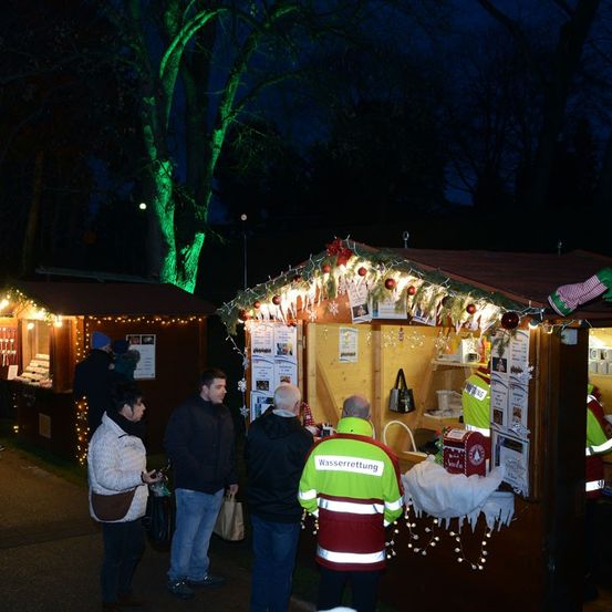 Menschen versammeln sich um einen festlich geschmückten Stand bei Nacht. Ein Mann in einer reflektierenden Weste steht drinnen, möglicherweise verkauft er Gegenstände. Ein anderer Mann hält eine Tasche, und eine Frau schaut zu. Die Szene wird von Weihnachtslichtern beleuchtet.