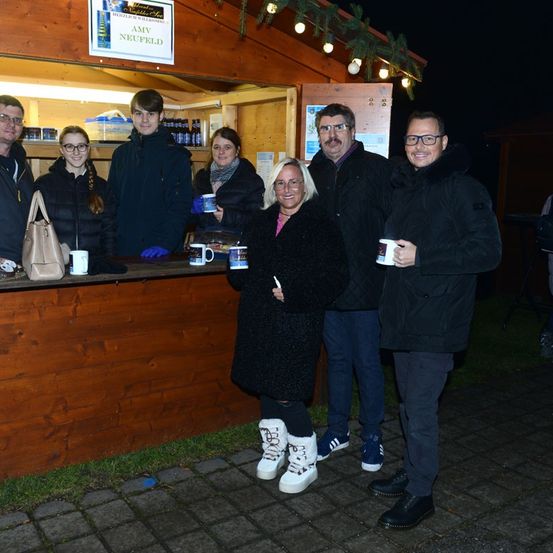Eine Gruppe von Menschen steht vor einem kleinen Holzhäuschen mit einem Schild, das 'AMV NEUFELD' besagt. Sie alle lächeln und halten Tassen in den Händen, wahrscheinlich für ein Foto posierend. Das Häuschen ist mit Weihnachtslichtern dekoriert.