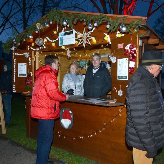 Ein Weihnachtsmarktstand mit Dekorationen und einem Mann in einer roten Jacke und einer Frau in einer grauen Jacke, die davor stehen.