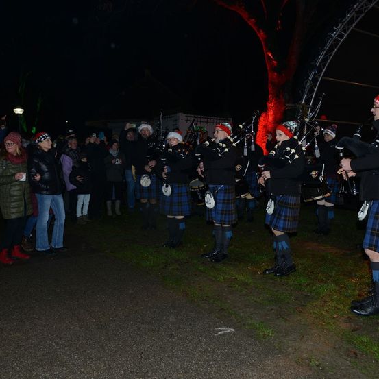 Eine Gruppe von Dudelsackpfeifer spielt bei Nacht vor einem Publikum unter roten Lichtern. Das Publikum ist warm gekleidet und einige machen Fotos.