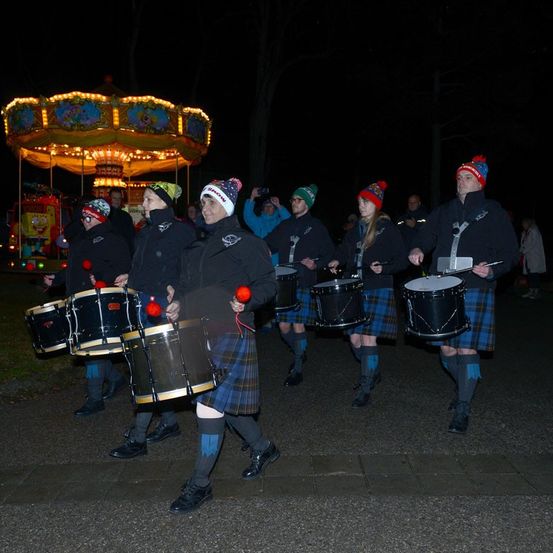 Eine Marschkapelle mit Trommeln und Dudelsack spielt bei Nacht vor einem beleuchteten Karussell.