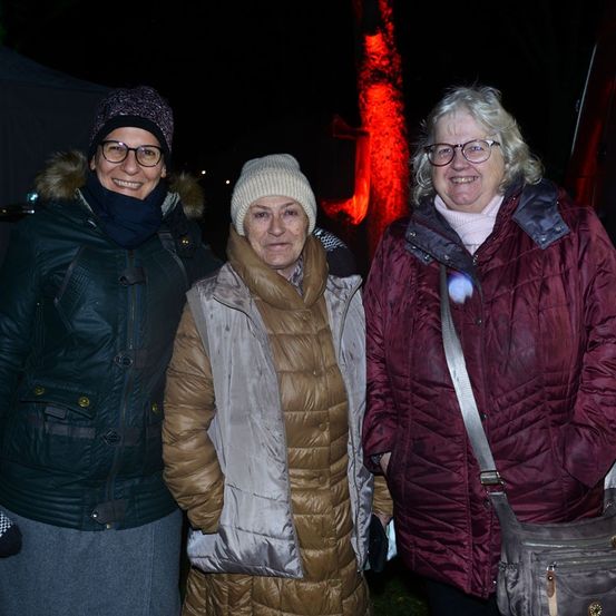 Drei Frauen, die für die kalte Nacht warm gekleidet sind, stehen zusammen. Sie lächeln und scheinen für ein Foto zu posieren. Die Frau auf der linken Seite trägt eine grüne Jacke und eine Mütze. Die Frau in der Mitte trägt einen braunen Mantel und eine weiße Mütze. Die Frau auf der rechten Seite trägt einen bordeauxfarbenen Mantel und hat eine Umhängetasche. Dahinter ist ein rotes Licht.