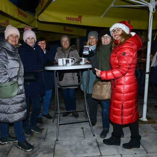 Eine Gruppe von Frauen steht im Freien unter Regenschirmen, eine hält eine Kaffeetasse. Sie tragen Winterkleidung, einschließlich Mützen und Handschuhen. Die Umgebung deutet auf ein gesellschaftliches Treffen oder eine Veranstaltung hin.