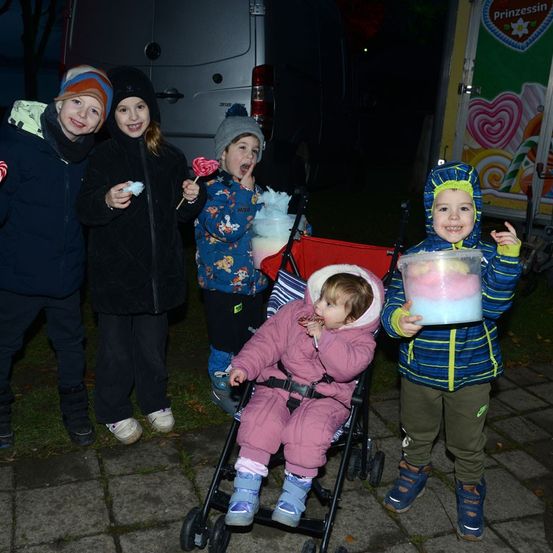 Eine Gruppe von Kindern, die lächeln und bei einer nächtlichen Veranstaltung Süßigkeiten genießen. Ein Baby im Kinderwagen, zwei Kinder mit Zuckerwatte und ein Kind mit einem Lollypop.