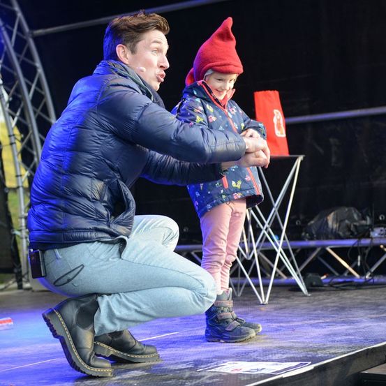 Ein Mann kniet auf der Bühne und hält die Hand eines Kindes, während ein Kind mit rotem Hut in der Nähe steht. Dahinter sind ein roter Beutel und Klappstühle zu sehen.