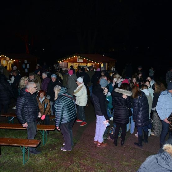 Eine Menschenmenge versammelt sich bei Nacht im Freien unter den Lichtern kleiner Stände. Die Leute tragen Winterkleidung und einige stehen, andere sitzen auf Bänken.