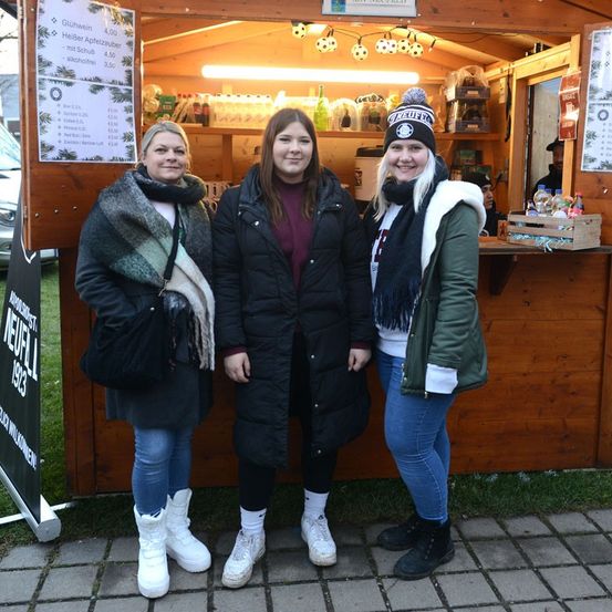 Drei Frauen in Winterkleidung stehen lächelnd vor einer Kamera vor einem Holzstand. Der Stand hat eine Preisliste und ausgestellte Artikel. Ein Auto ist hinter ihnen geparkt.