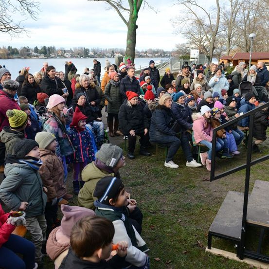 Eine Menschenmenge, bestehend aus Erwachsenen und Kindern, ist in einem Außenbereich versammelt. Einige sitzen auf dem Gras, während andere stehen. Einige von ihnen tragen Winterkleidung, Mützen und Handschuhe. Vor ihnen befindet sich eine Treppe mit Handlauf. In der Ferne gibt es einen See mit einem Boot und Bäumen.
