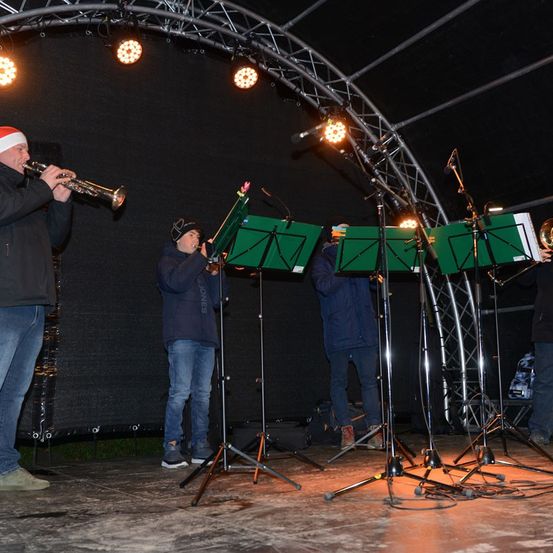 Eine Gruppe von Menschen spielt auf einer Bühne mit Trompeten. Die Bühne hat einen schwarzen Hintergrund und Metallstrukturen. Sie tragen Winterkleidung und einige von ihnen tragen Mützen. Sie spielen, während sie vor einem Mikrofon stehen.