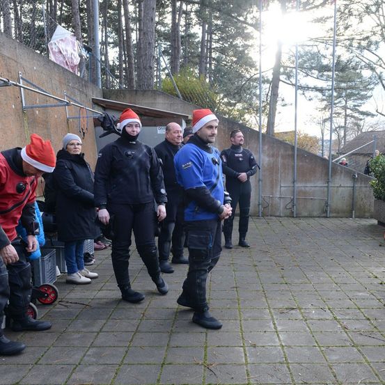 Eine Gruppe von Menschen in Taucheranzügen und Weihnachtsmützen steht auf einem gepflasterten Weg. Sie scheinen sich auf ein Tauchen vorzubereiten. Im Hintergrund befinden sich Bäume und ein Gebäude.