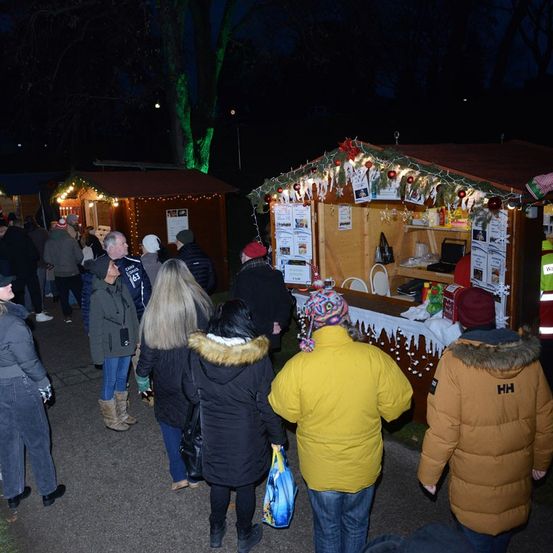 Eine Menschenmenge versammelt sich bei Nacht um beleuchtete Weihnachtsstände. Die Stände sind mit Lichtern und Weihnachtsdekorationen geschmückt. Die Leute stöbern durch die Stände, einige tragen Winterkleidung.