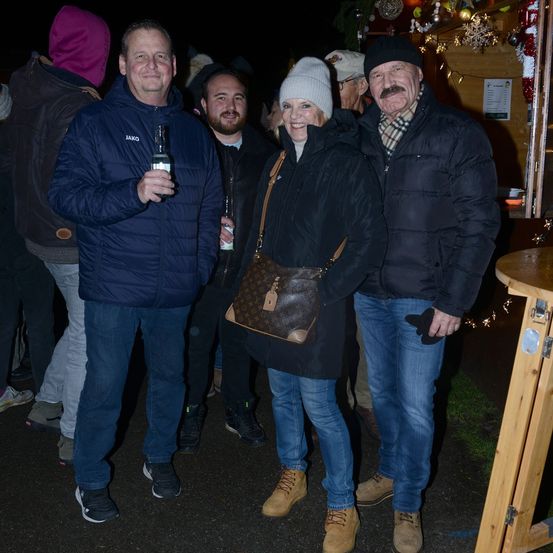 Drei Erwachsene stehen bei Nacht im Freien. Ein Mann hält eine Flasche und zwei Frauen stehen neben ihm. Dahinter stehen weitere Personen, von denen einige verschwommen sind.