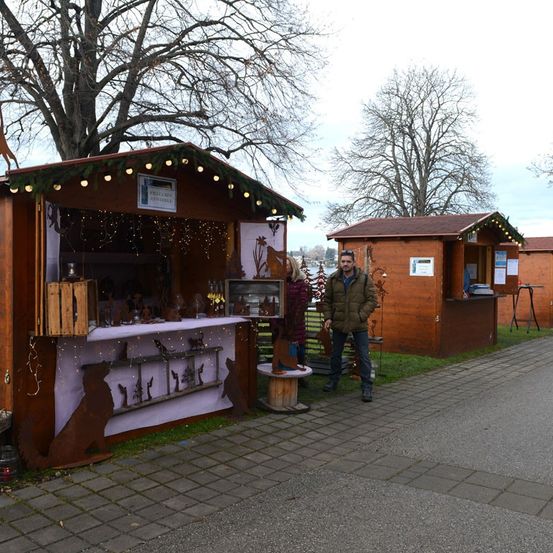 Ein Mann und eine Frau stehen neben zwei Holzhütten auf einem gepflasterten Weg. Die Hütten sind mit Lichtern und einem Weihnachtsbaum dekoriert. Eine Hundesilhouette schmückt die Vorderseite einer Hütte.