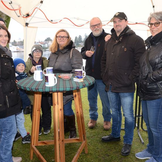 Eine Gruppe von Menschen steht um einen kleinen Tisch unter einem Zelt. Drei Tassen stehen auf dem Tisch. Zwei Männer tragen Jacken und einer trägt einen Hut. Dahinter befindet sich ein See und eine verschwommene Ansicht von Bäumen und Häusern.