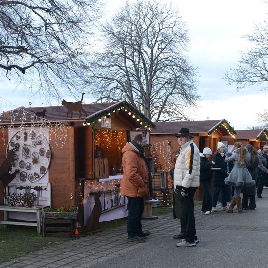 Eine Gruppe von Menschen besucht einen Weihnachtsmarkt. Sie stehen vor hölzernen Ständen mit festlichen Dekorationen. Bäume ohne Blätter und kahle Äste umgeben das Gebiet.