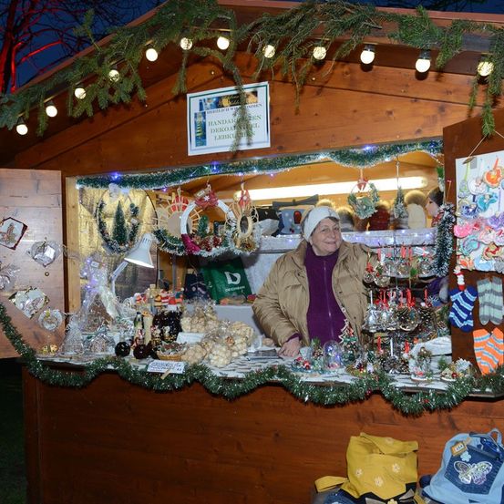 Eine lächelnde Frau steht hinter einem festlich dekorierten Holzstand und verkauft handgefertigte Weihnachtsdekorationen und Ornamente. Der Stand ist mit Weihnachtslichtern, Girlanden und verschiedenen dekorativen Gegenständen geschmückt.