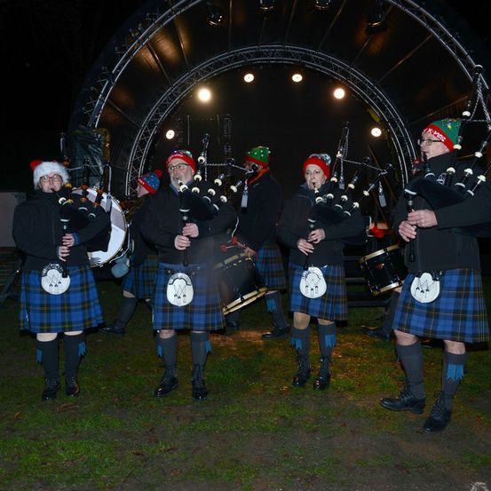 Eine Dudelsackband in Kilts tritt bei Nacht auf einer Bühne mit einem Metallbogen auf, beleuchtet von Scheinwerfern.