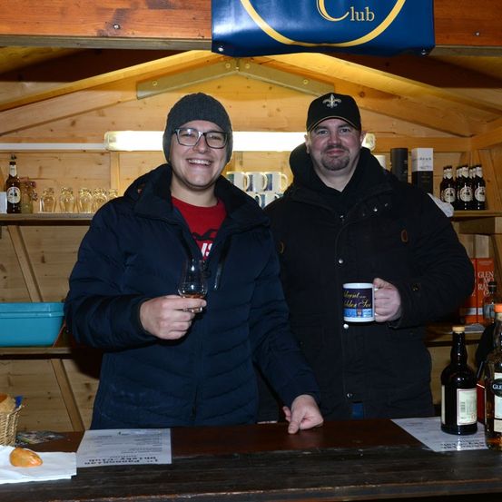 Zwei Männer stehen lächelnd hinter einer Holzbar und halten Getränke in den Händen. Der Mann auf der linken Seite hält ein Weinglas, während der Mann auf der rechten Seite einen Kaffeebecher hält. Dahinter befindet sich ein Holzregal mit Flaschen und Gläsern. Das Schild darüber zeigt 'Club.'
