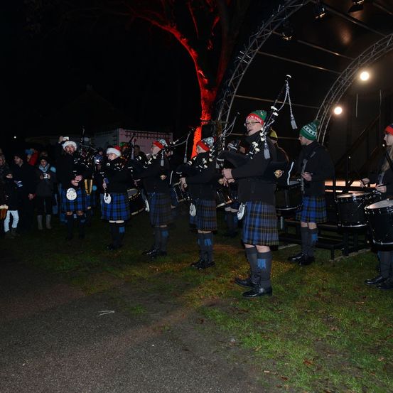 Eine Dudelsackband tritt nachts auf, gekleidet in Kilts und Mützen. Sie spielen Dudelsack und Trommeln vor einem Publikum. Der Bereich wird von roten Lichtern beleuchtet.