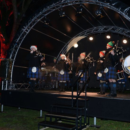Eine Gruppe von Musikern in Kilts tritt bei Nacht auf einer Bühne auf. Sie spielen Dudelsack, Trommeln und andere Instrumente, mit Scheinwerfern, die die Szene beleuchten.