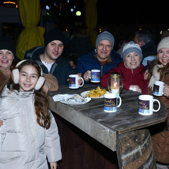 Eine Familie sitzt um einen Tisch herum, lächelt und posiert für ein Foto. Sie tragen Winterkleidung, und auf dem Tisch stehen Tassen und Teller.