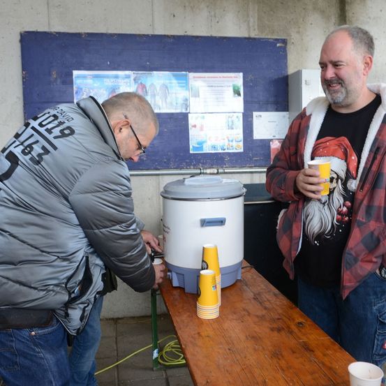 Zwei Männer stehen in einem Raum. Ein Mann gießt ein Getränk in einen Becher. Der andere Mann hält einen Becher und lächelt. Es gibt Becher und einen Behälter auf einem Holztisch. Es gibt Plakate an der Wand.